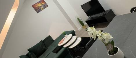 Una sala de estar moderna con un sofá verde, un televisor de pantalla plana, una mesa de comedor con un jarrón de flores y un reloj en la pared.
