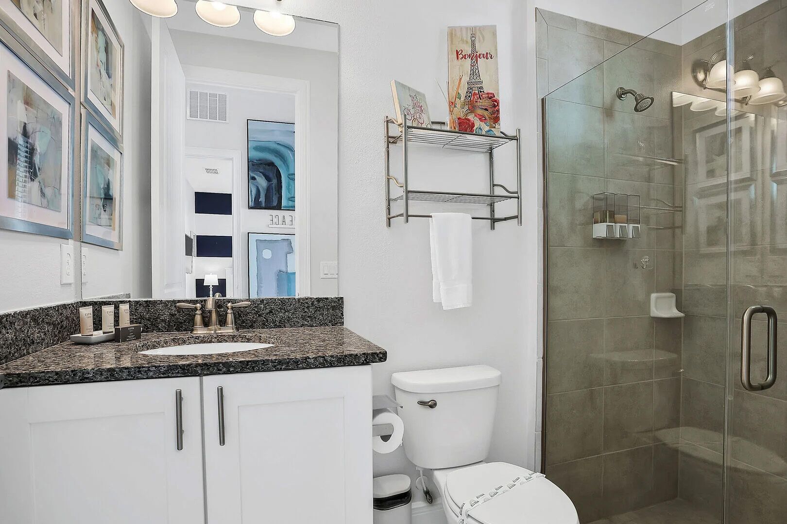 A modern bathroom with a granite countertop, white fixtures, a glass shower enclosure, and artwork on the walls.