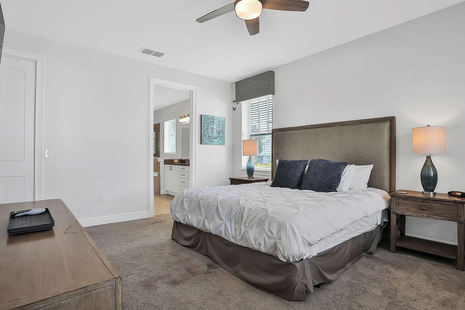 Une chambre à coucher comprenant un lit, des tables de chevet, un ventilateur de plafond, un miroir et une porte donnant sur une salle de bain.