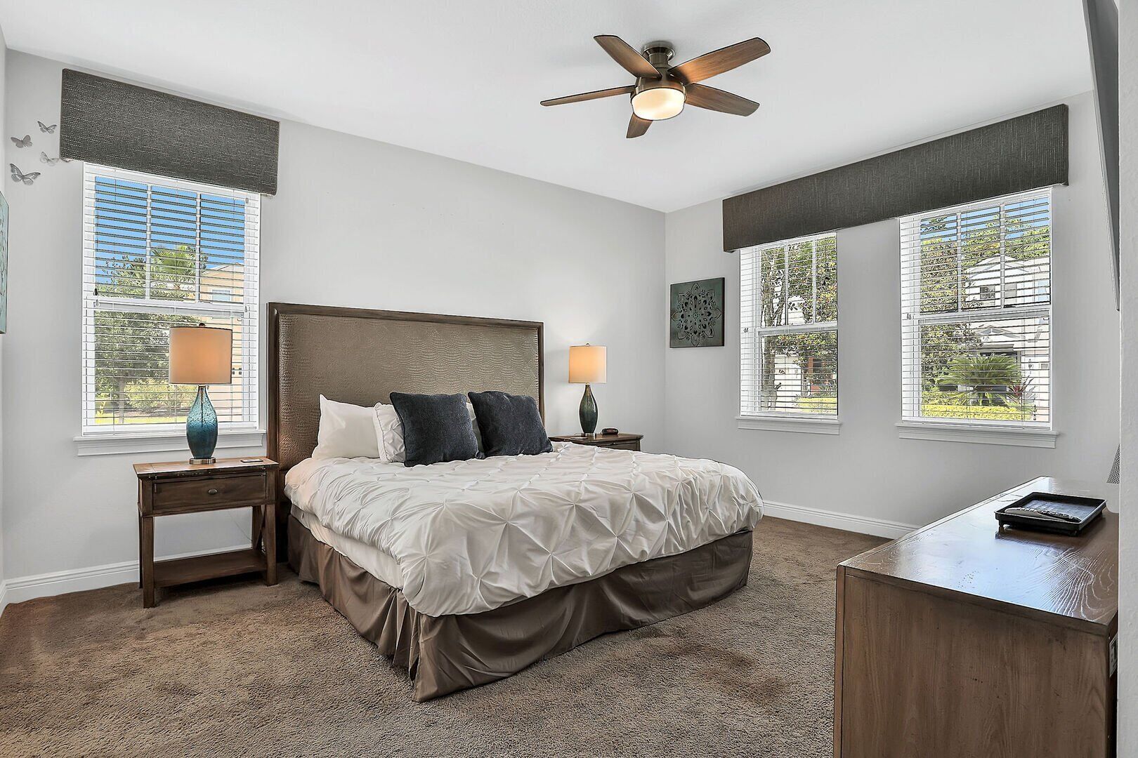 A bedroom with a bed, two bedside tables, a desk, a ceiling fan, and windows with blinds.