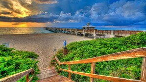 A wooden pier leading to a beach with a lifeguard tower, surrounded by greenery.