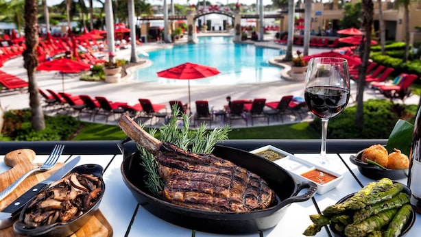 A dining table with a grilled steak, asparagus, and a glass of red wine, overlooking a pool area with red umbrellas and lounge chairs.