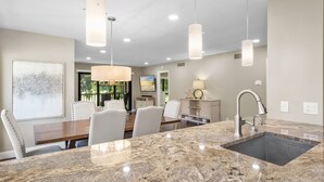 A modern kitchen with granite countertops, a built-in sink, pendant lighting, and a dining area with white chairs and a wooden table.