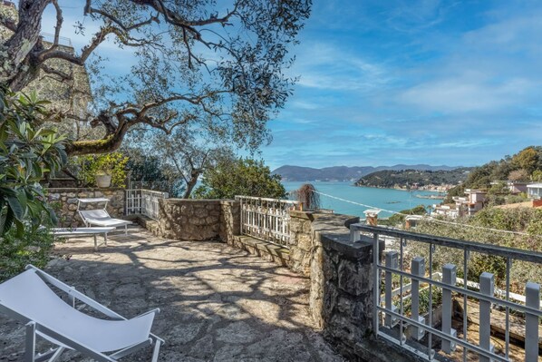 A stone patio with lounge chairs, a stone wall, and a view of a coastal town and mountains.
