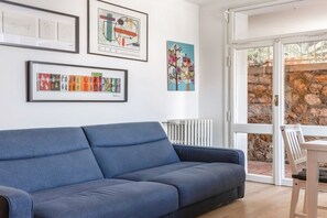 A modern living room with a blue sofa, framed artwork on the wall, and a stone wall visible through the glass door. - Comfortable apartment perfect for 4 people and surrounded by greenery, it is located in a quiet area, a few steps from the beaches of Lerici. The house is equipped with every comfort, 2 bathrooms, kitchen, TV, WiFi, private parking. Ideal opportunity to  (Lerici)