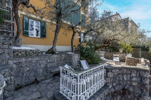 A stone staircase leading to a yellow building with green shutters, a white metal railing, and a tree in the foreground.