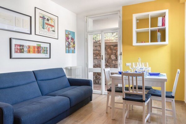 A room with a blue sofa, a dining table with chairs, a window with a view, and framed artwork on the wall.
