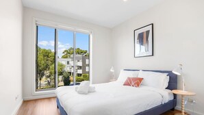 Una habitación de hotel moderna con una cama grande, una mesita de noche, una lámpara de pie y un ventanal con vistas a la ciudad.