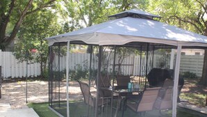 Un patio techado con mesa y sillas, rodeado por una valla blanca y árboles.