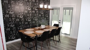 Un área de comedor con un muro temático de café en blanco y negro, una mesa de madera con individuales, sillas negras y una lámpara colgante.