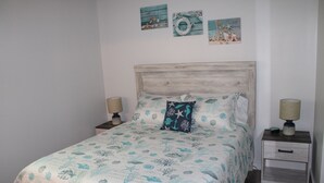Un dormitorio con cabecera de madera, cama con cubrecama de temática marina en azul y blanco, dos mesitas de noche con lámparas, y tres cuadros en la pared sobre la cama.