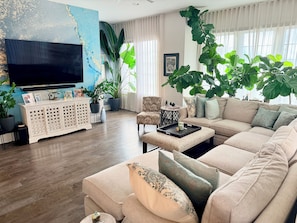 A living room with a sofa, coffee table, and a TV. There are potted plants and a cabinet with photos.