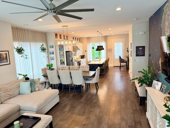 A spacious living area with a sofa, dining table, and chairs, a kitchen with a refrigerator, and a ceiling fan.