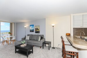 Ein modernes Wohnzimmer mit einem grauen Sofa, einem Couchtisch und einem Essbereich mit einem Stuhl und einem kleinen Tisch. Zwei Stehlampen und Kunstwerke sind an der Wand angebracht.