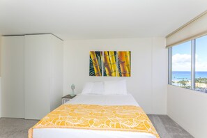 Ein Hotelzimmer mit Bett, Nachttisch, Lampe, Fenster mit Blick auf den Strand und einem Bambusgemälde.