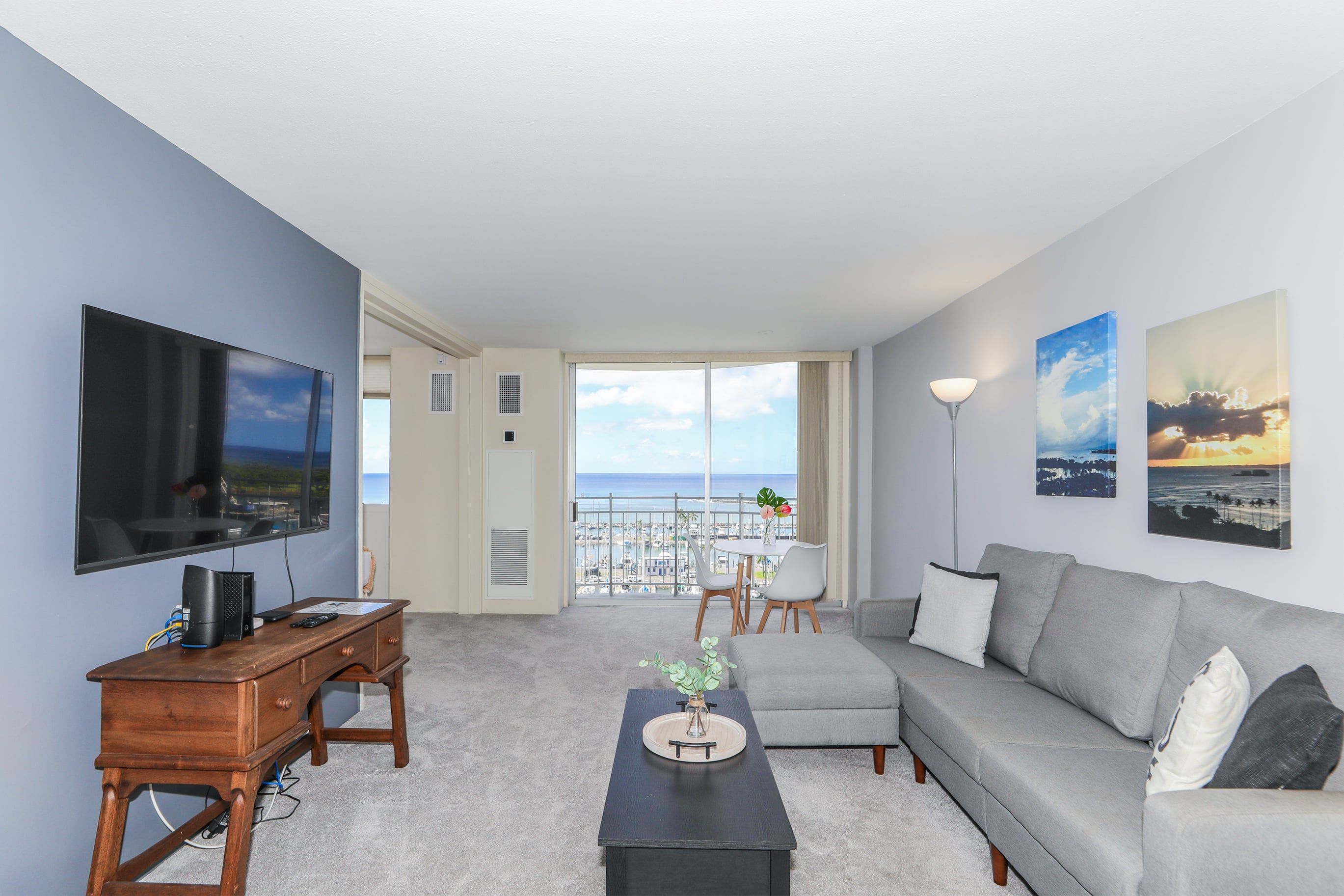 A modern living room with a grey sofa, a coffee table, and a TV on a wooden stand. There is a balcony with a view of the sea and a dining area with chairs and a table.