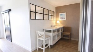 Een kleine, moderne kamer met een wit bureau, een stoel, een lamp en een ingelijst schilderij aan de muur.