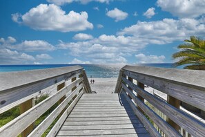 Une promenade en bois menant à une plage, avec un ciel dégagé et un palmier.