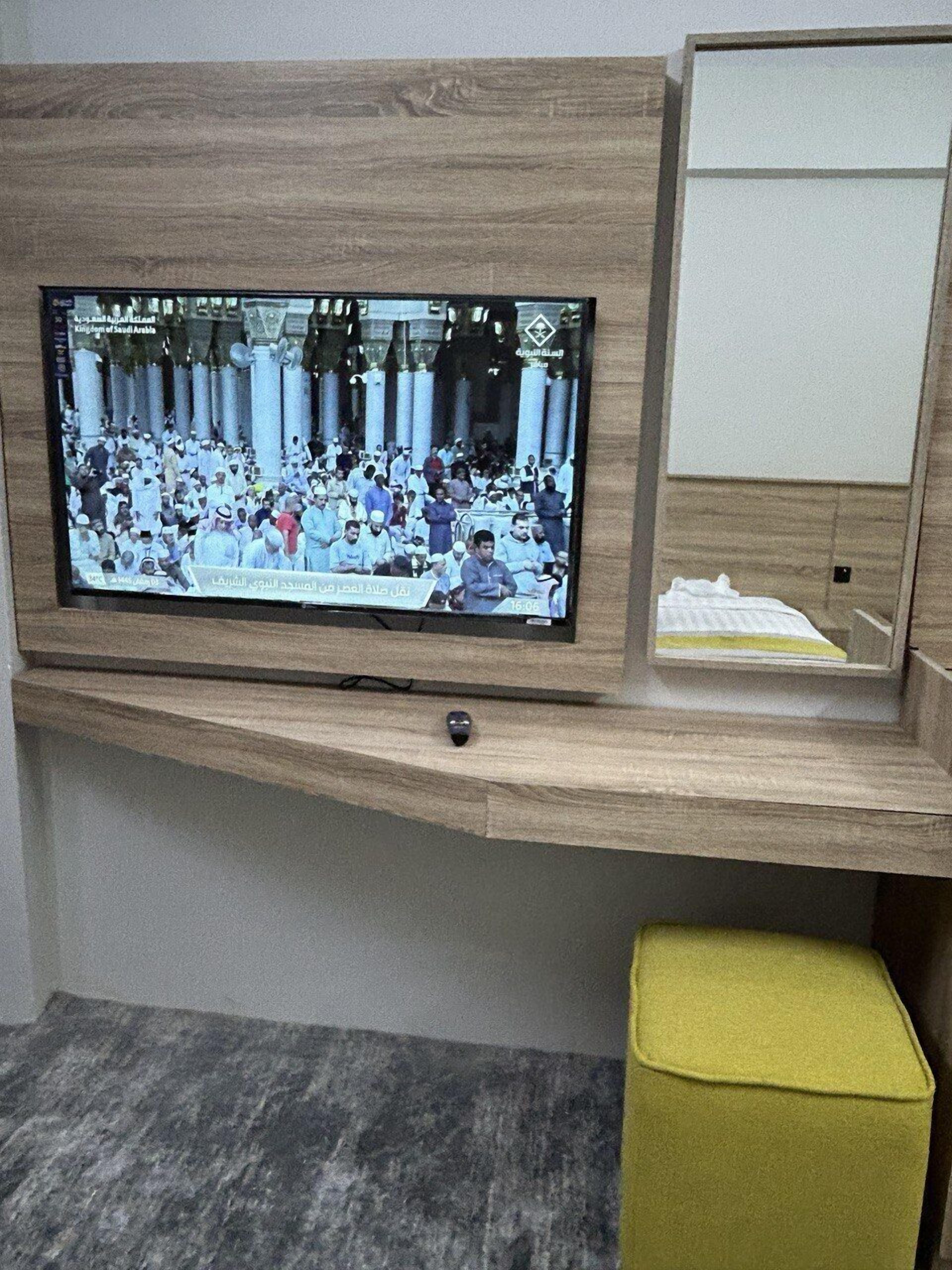 A television mounted on a wooden wall, a small yellow stool, and a cubicle-style desk with a white surface.