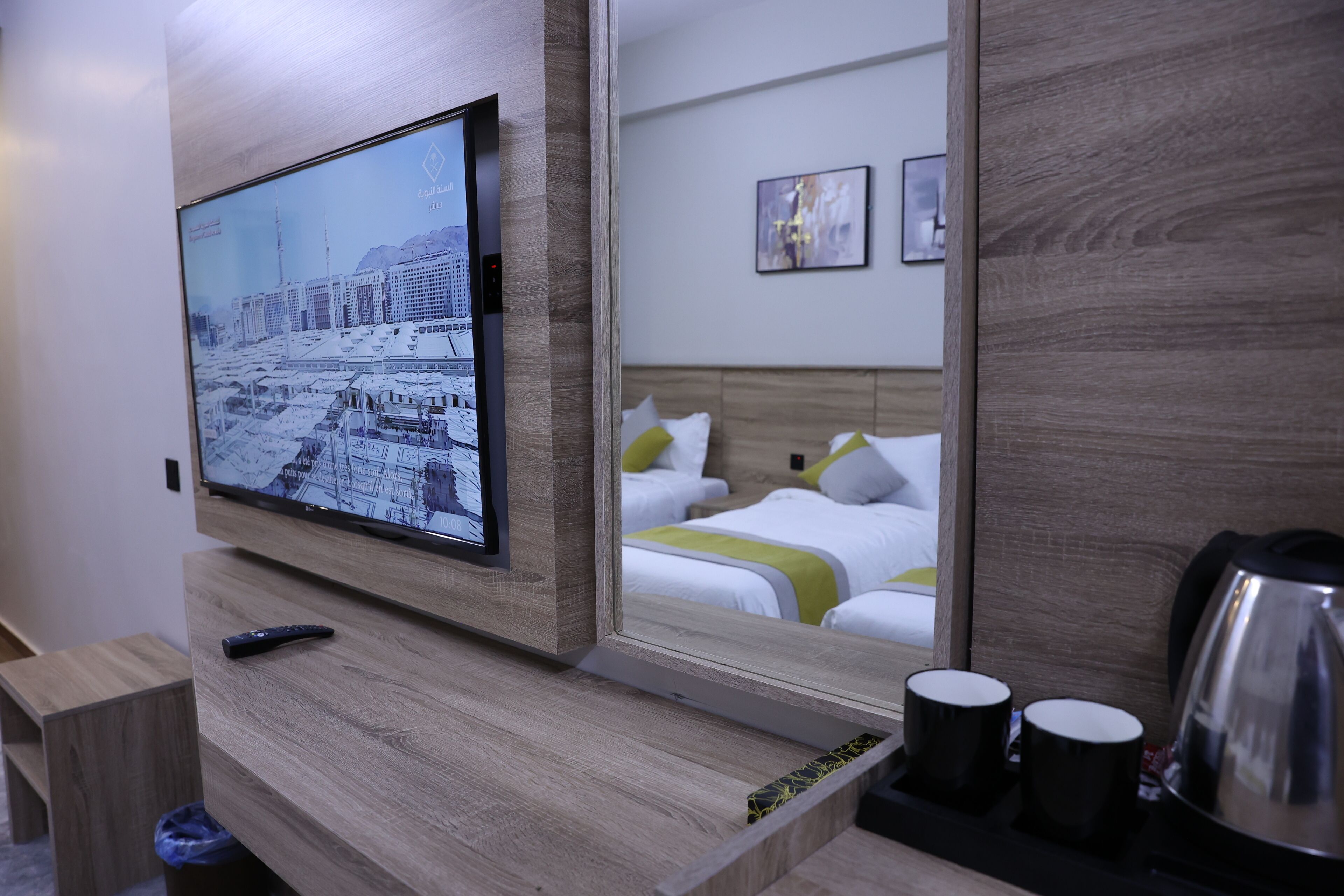 A hotel room with a mounted TV displaying a cityscape, a bed with white bedding and green accents, a bedside table with a remote control, and a coffee maker.
