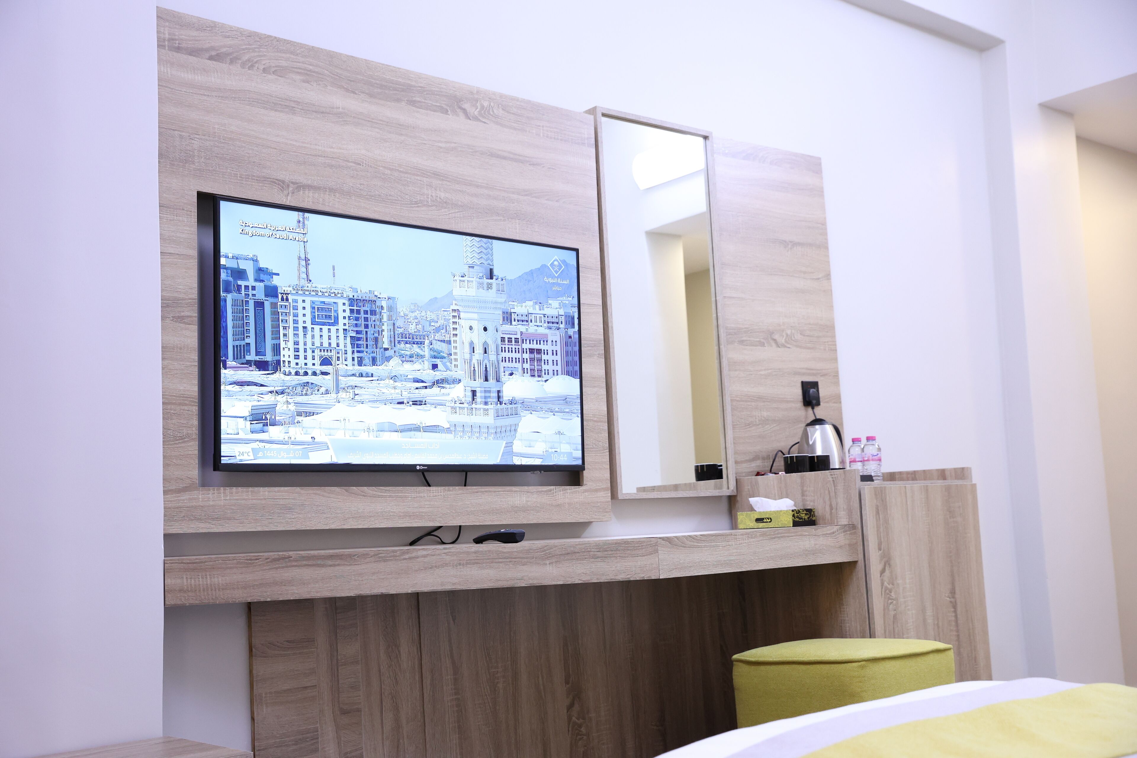 A modern hotel room with a large flat-screen TV mounted on a wooden wall, a bedside table with a lamp, and a green ottoman.