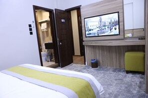Una habitación de hotel con una cama, un televisor montado en la pared, un escritorio de madera, un otomano verde y un baño visible a través de una puerta abierta.