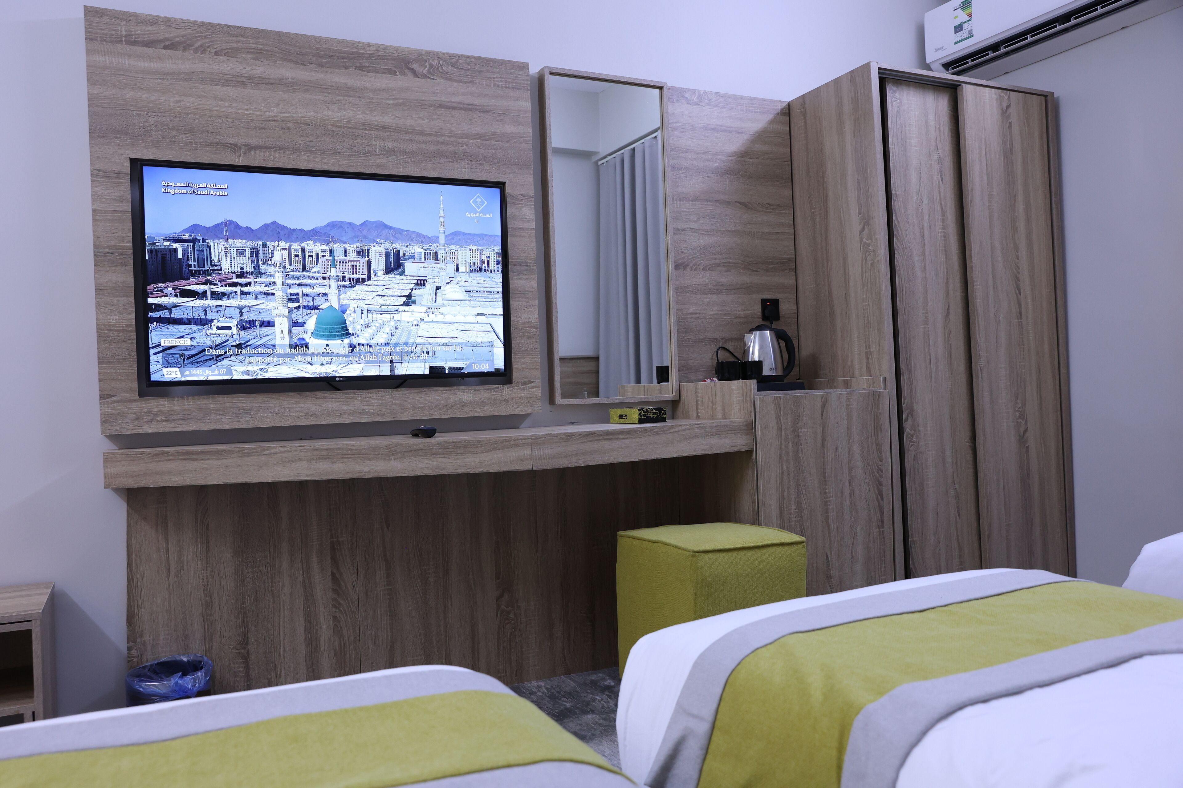 A hotel room with a television mounted on the wall, a wardrobe, a bed with green and white bedding, and a small stool.