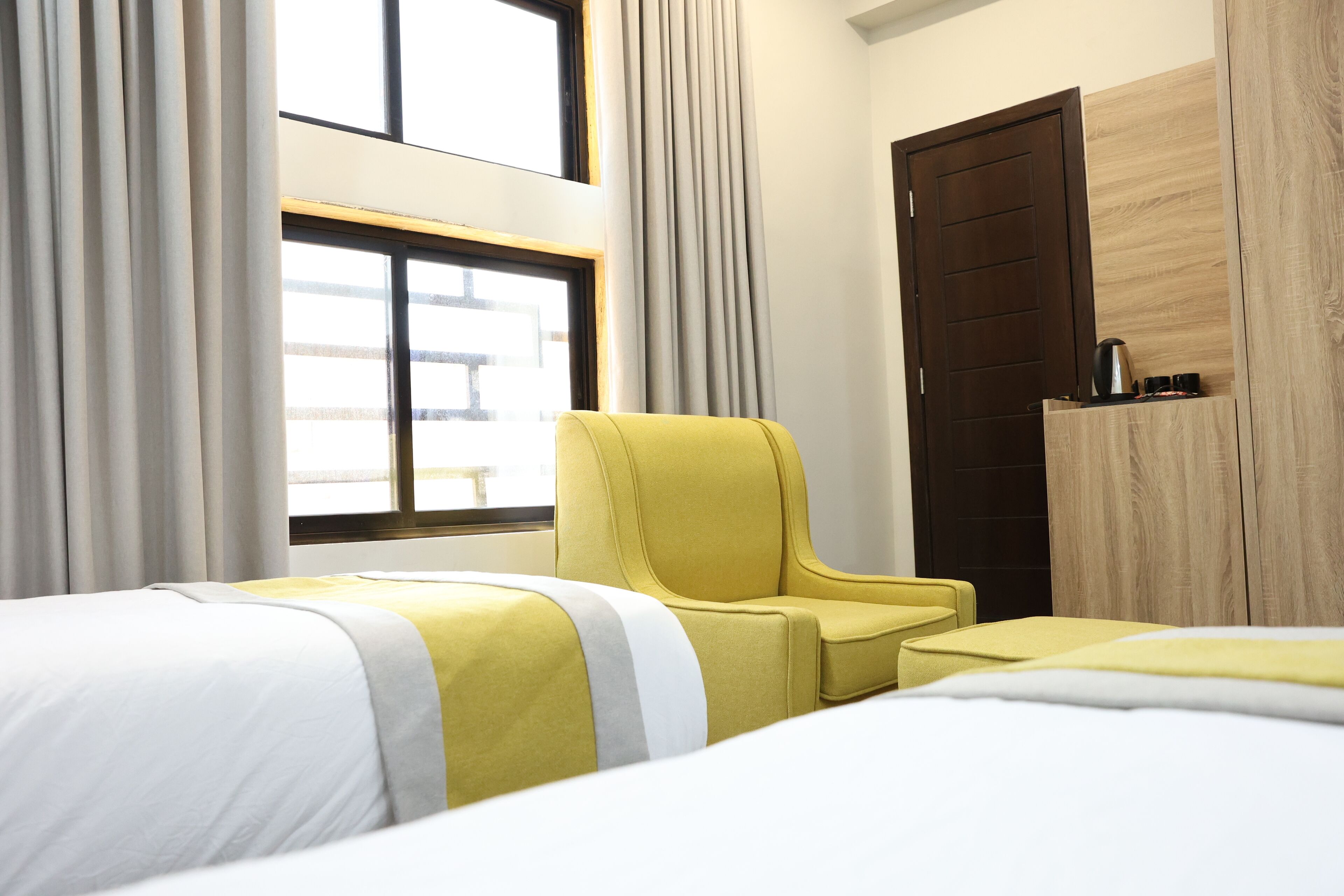 A hotel room with two beds, a yellow armchair, a wooden wardrobe, and a window with curtains.