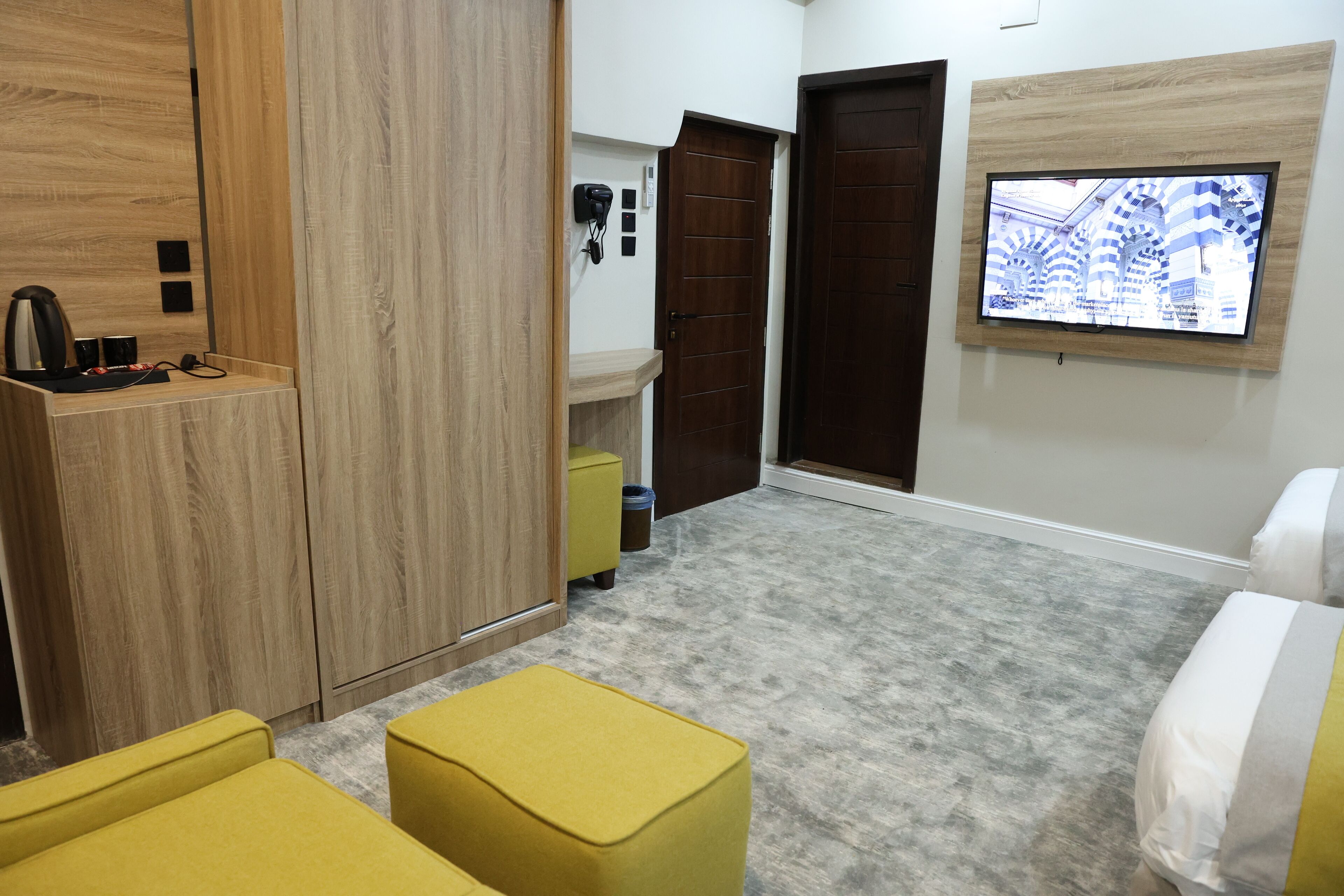 Habitación de hotel con un armario de madera, un pequeño escritorio con hervidor, un taburete amarillo, un televisor montado en la pared y un ventanal grande.