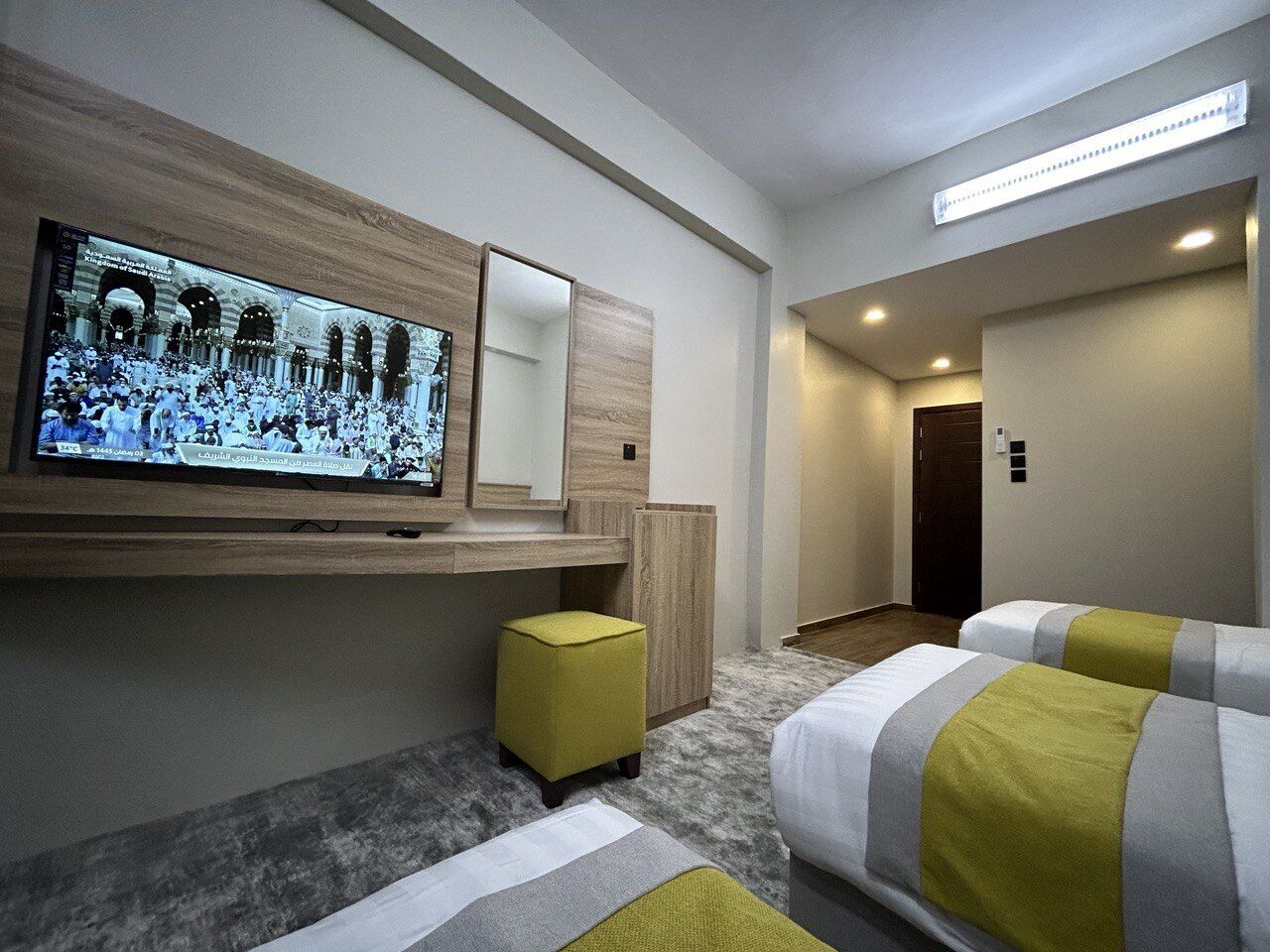Een hotelkamer met een aan de muur bevestigde televisie, een klein geel poufje en twee bedden met gestreepte beddengoed.