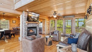Un intérieur de chalet chaleureux avec une cheminée en pierre, des meubles en cuir et de grandes fenêtres offrant une vue sur l’extérieur.