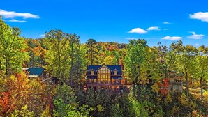 Une grande cabane en rondins à deux étages, dotée d’une fenêtre centrale imposante et d’une véranda qui fait le tour de la maison, le tout entouré d’une forêt dense.