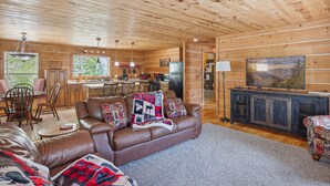 A cozy cabin interior with a brown leather sofa, a dining table with chairs, a kitchen area, and a large window overlooking greenery. - The "Lost Horse" Cabin - Secluded - Pet-Friendly - One Level - Massive Yard (Sevierville)