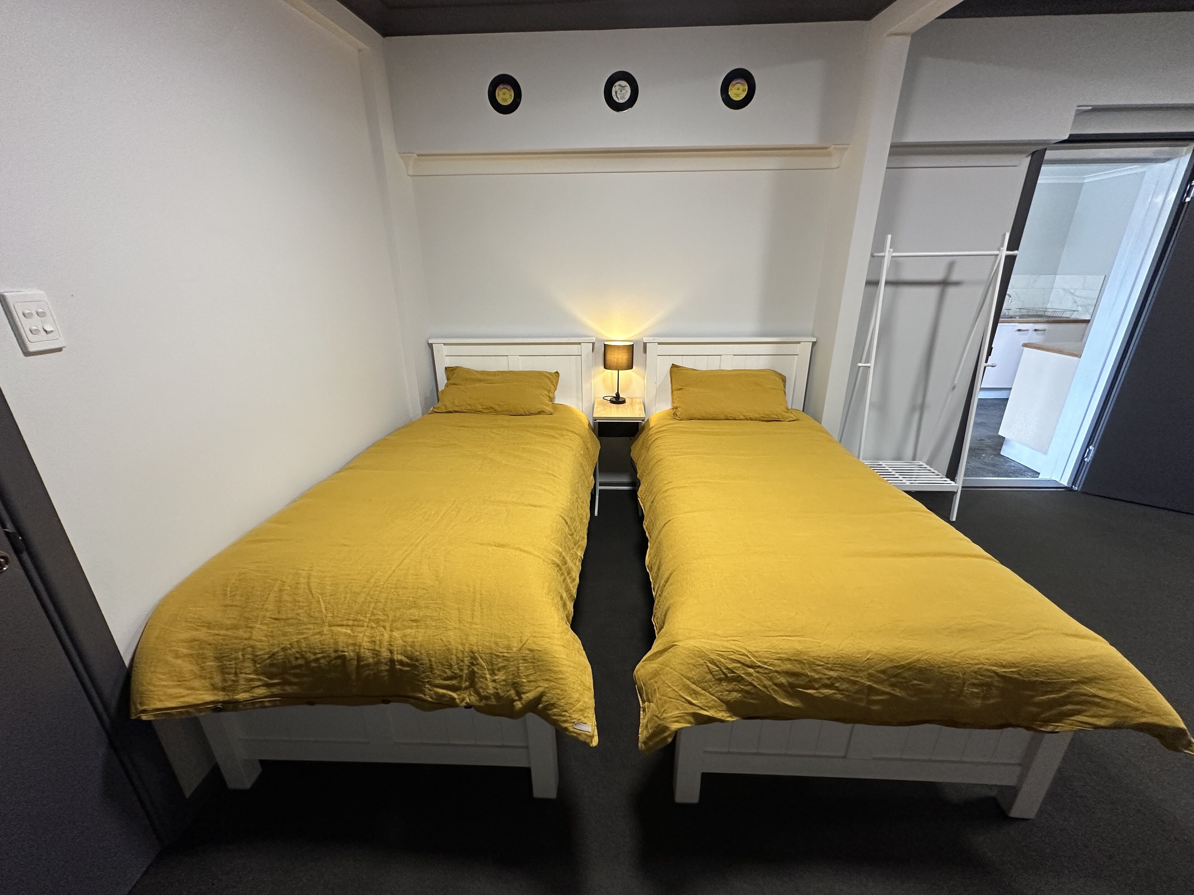 Een kamer met twee bedden, elk met een gele sprei, een nachtkastje met een lamp, en een wandplank met drie ronde objecten.