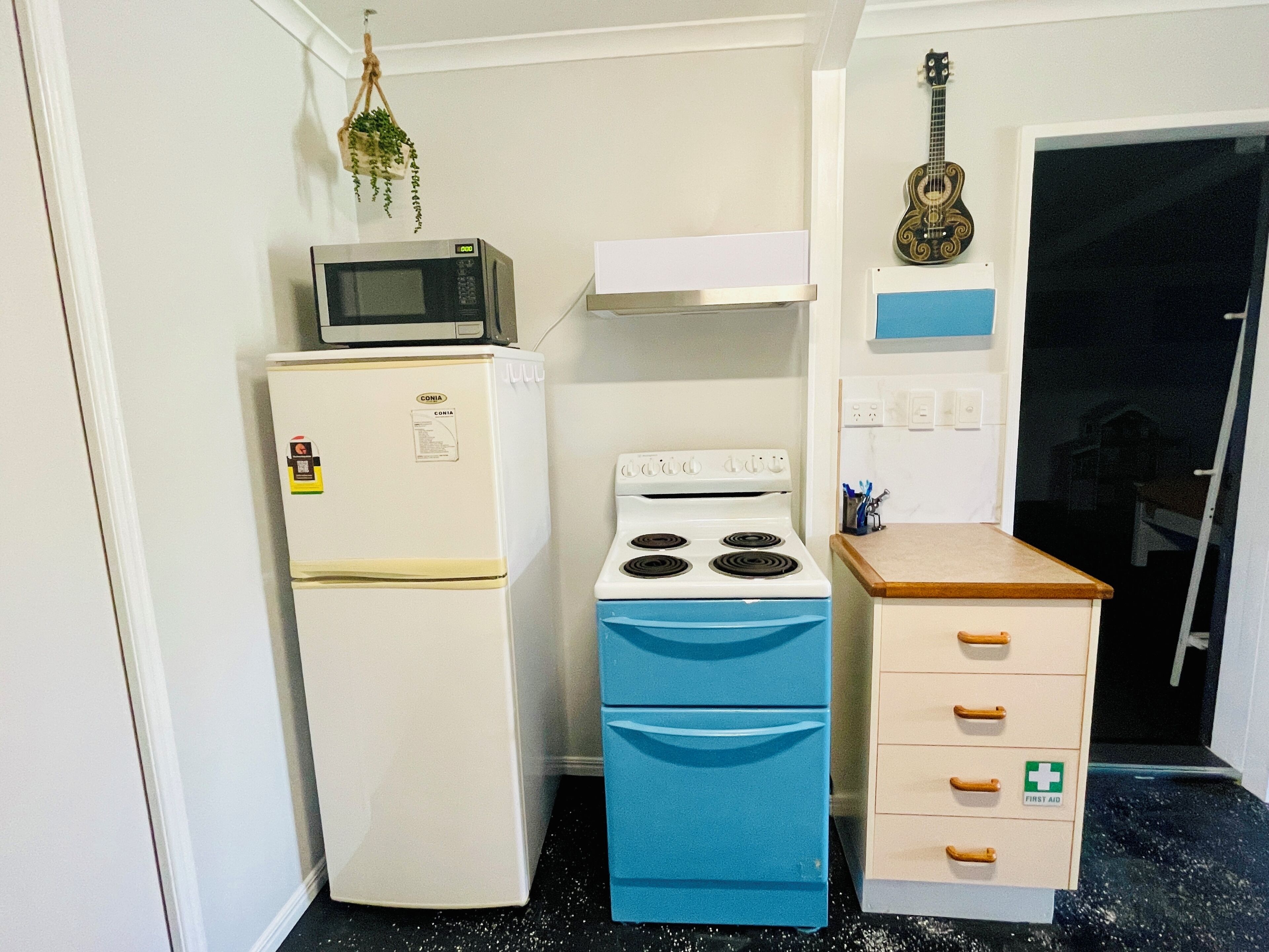 Une cuisine de taille réduite avec un réfrigérateur blanc, une cuisinière bleue et un four à micro-ondes posé sur le réfrigérateur. On y trouve un petit placard avec un panneau indiquant la présence d’une trousse de premiers secours, et une guitare accrochée au mur.