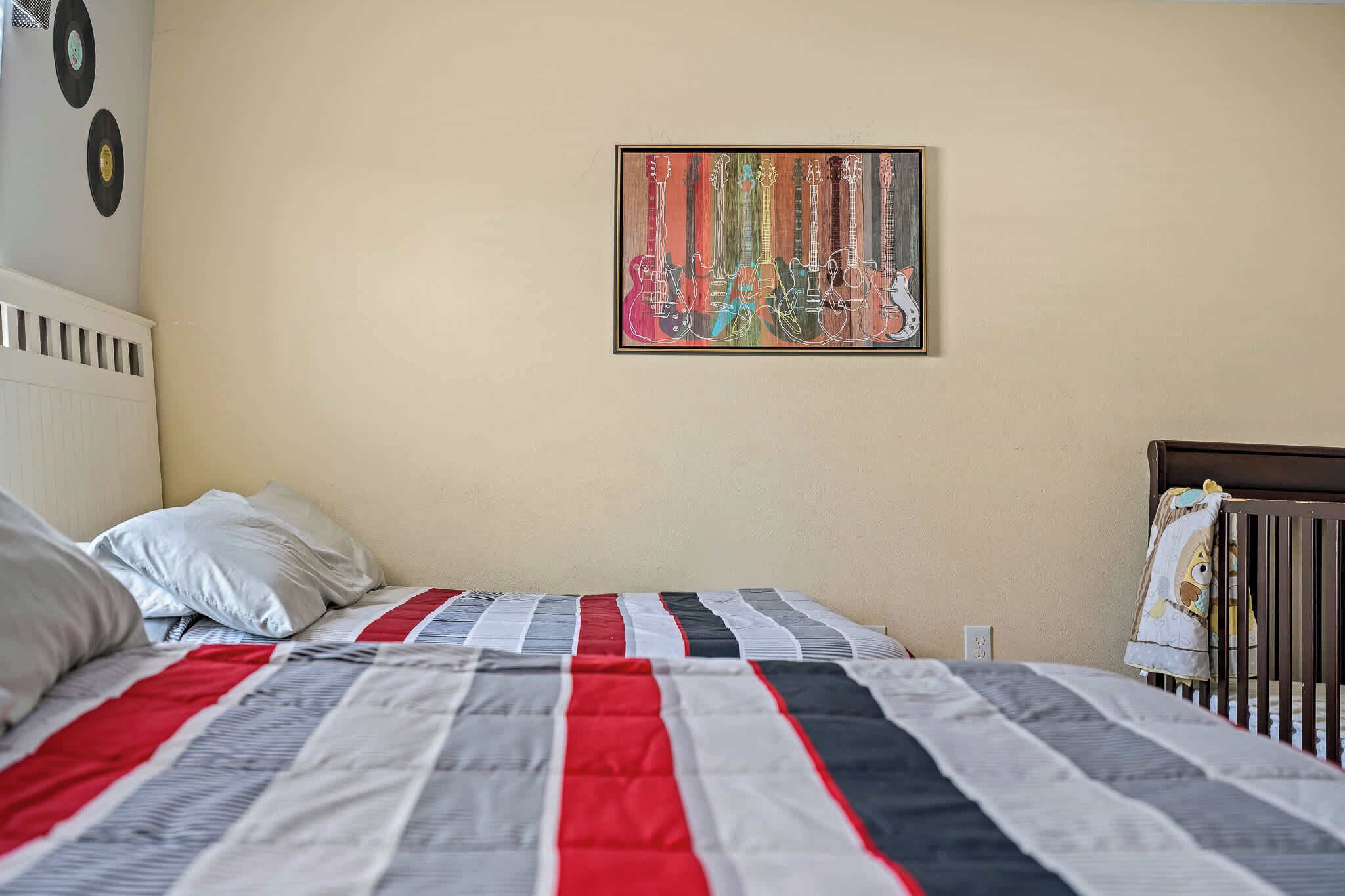 A bedroom with a bed, pillows, a crib, and a wall with a framed picture.