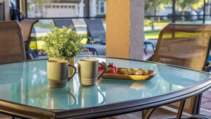 Een glazen tafel met twee mokken en een dienblad eten, een kamerplant, en terras met uitzicht op huizen en bomen.