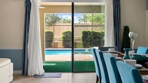Ein Zimmer mit einer Schiebetür zu einem Poolbereich, einem Esstisch mit blauen Stühlen und einem kleinen Beistelltisch mit einem dekorativen Gegenstand.