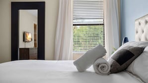 Ein ordentlich bezogenes Bett mit aufgerollten Handtüchern und Kissen, ein großer Spiegel an der Wand und ein Fenster mit Jalousien.