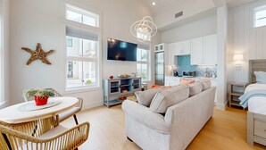 A spacious living area with a grey sofa, a dining table with wicker chairs, a TV mounted on the wall, and a kitchen area with white cabinets and a blue backsplash. - Resort Style Pools, Golf Cart Boardwalk to the Beach (Port Aransas)