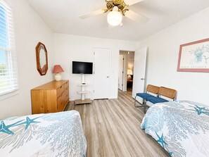 Une chambre avec deux lits, une télévision, un ventilateur de plafond et une porte donnant sur une autre pièce.