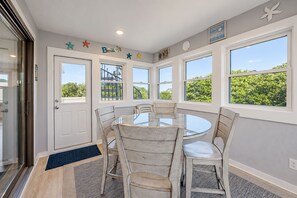Un espace repas comprenant une table ronde et six chaises, de grandes fenêtres donnant sur un paysage verdoyant, et une porte dotée d’un panneau en verre.