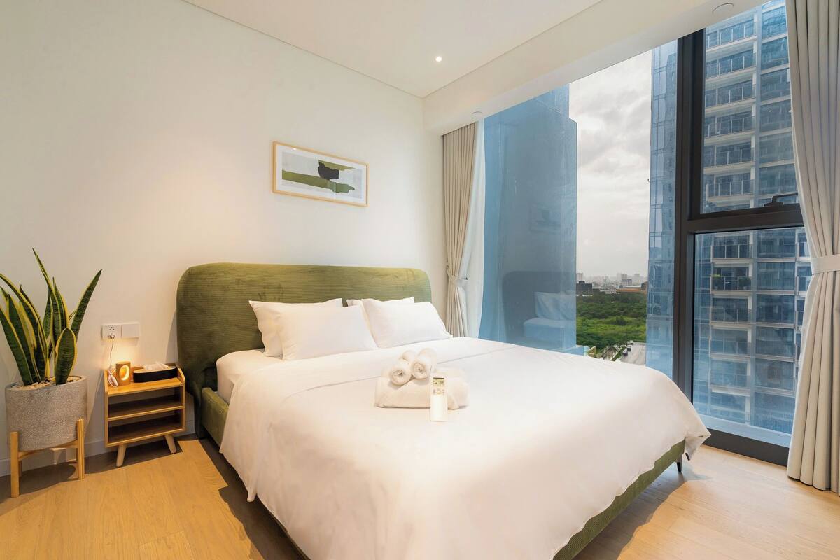 Une chambre d’hôtel moderne avec un grand lit, une table de chevet avec une lampe, une plante en pot et une vue sur la ville grâce à de grandes fenêtres.