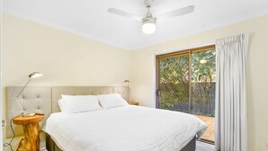 A bedroom with a bed, a ceiling fan, a nightstand with a lamp, and a view of a garden through a sliding glass door.