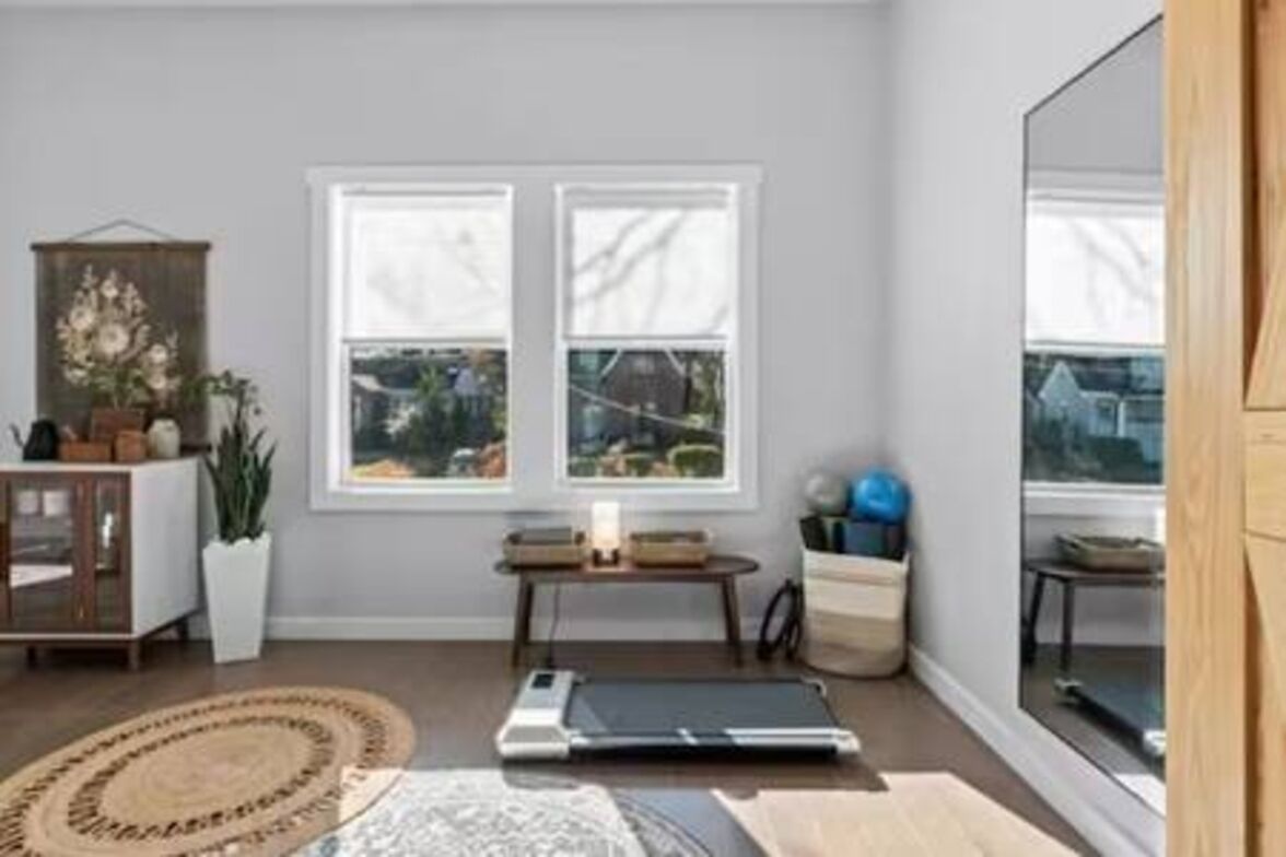 Ein Zimmer mit großem Fenster, Laufband, Schrank und Pflanze.