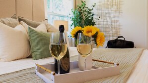 Un lit avec un plateau contenant une bouteille de vin et deux verres à vin, un vase avec des tournesols et un sac noir posé à côté.