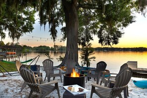 Un espace extérieur aménagé au bord du lac, avec des chaises Adirondack, un foyer et une vue sur le lac au coucher du soleil.