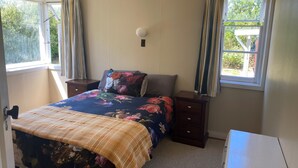 A bedroom with a bed, a bedside table, a nightstand, a lamp, and a window with curtains. - Our Bach. Fish, ski or relax in an amazing alpine setting! (Lake Coleridge)