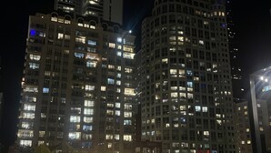 Nachtsicht auf einen Hochhauskomplex mit beleuchteten Fenstern, einer Feuerstelle im Vordergrund und Außenbestuhlung.