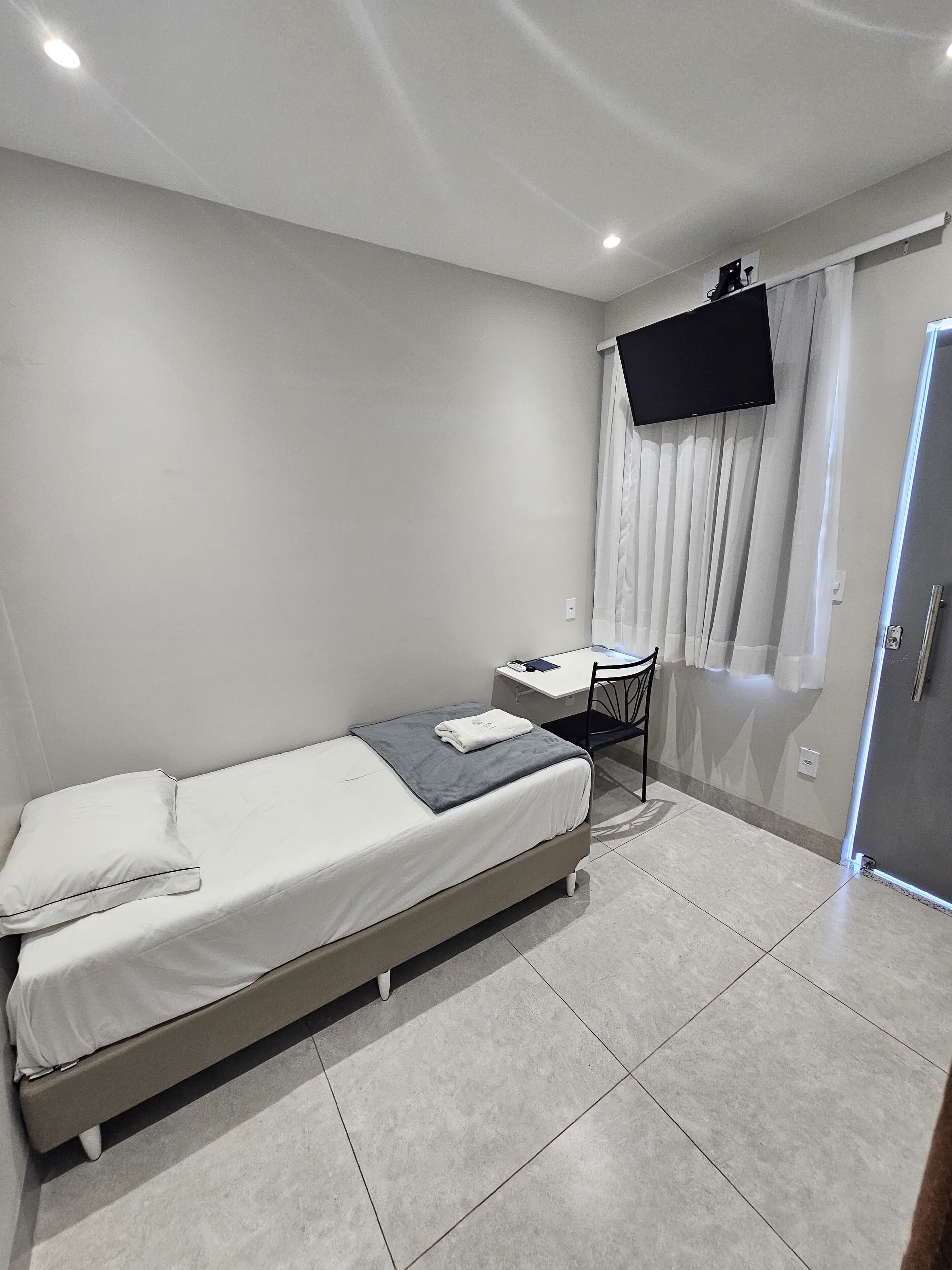 A small, modern bedroom with a single bed, a desk, and a TV mounted on the wall.